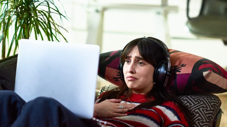 Eine Person liegt auf einem Sofa, trägt Kopfhörer und schaut etwas auf einem Laptop.