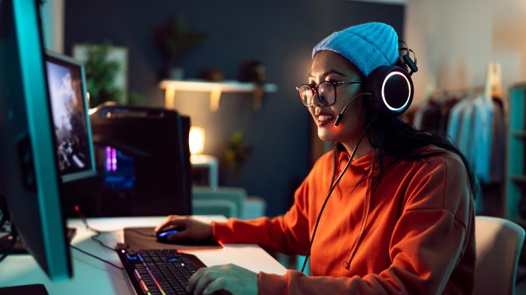 Eine Person mit Headset auf dem Kopf sitzt an einem Schreibstisch vor zwei Monitoren neben einem Gaming-PC.