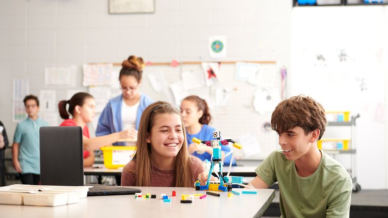 Zwei Kinder bauen einen Roboter mit LEGO Spike. Im Hintergrund sind weitere Kinder zu sehen.
