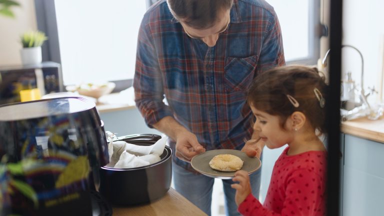 Eine Person steht mit einem Kind vor einem geöffneten Airfryer und begutachtet einen Donut auf einem Teller.