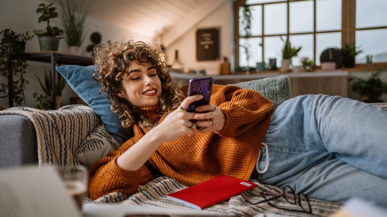 Eine Person liegt auf der Couch im Wohnzimmer mit Pflanzen im Hintergrund und scrollt auf ihrem Smartphone.