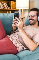 Eine Person liegt auf einem Sofa, trägt Kopfhörer und hält ein Smartphone in der Hand.