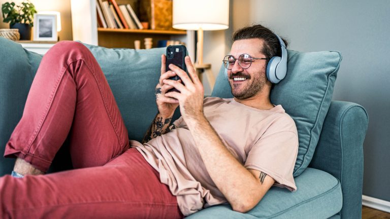 Eine Person liegt auf einem Sofa, trägt Kopfhörer und hält ein Smartphone in der Hand.