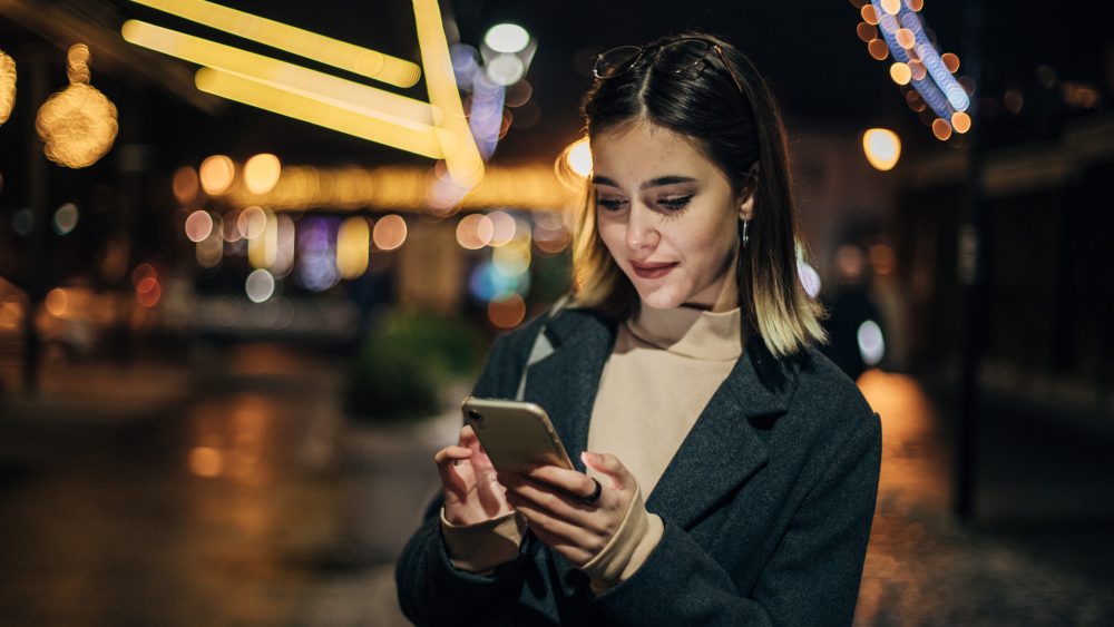 Eine Person steht nachts auf der Straße und blickt auf ihr Smartphone.