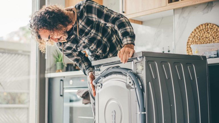 Eine Person beugt sich über eine Waschmaschine, um deren Hinterseite zu begutachten.