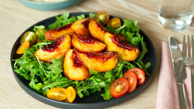 Ein Salat mit Pfirsich, Ruccola und Tomaten steht auf einem Tisch.