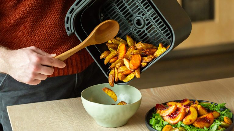 Eine Person gießt einige Kartoffelspalten von einem Airfryer-Korb in eine Schüssel.