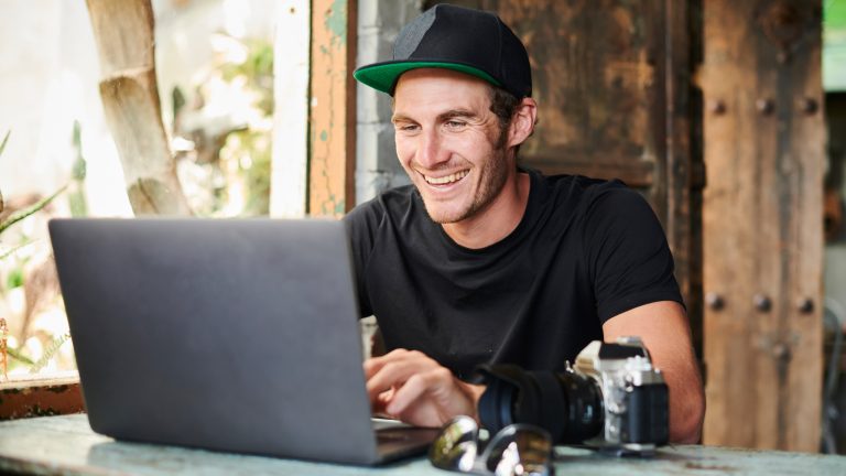 Eine Person mit einer Kamera neben ihr auf dem Tisch sitzt an einem Laptop und grinst.