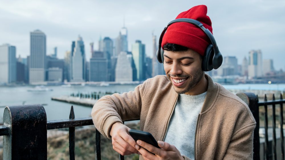 Eine Person steht vor einem Stadtpanorama und schaut auf ihr Handy, während sie über Headphones Musik hört.