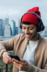 Eine Person steht vor einem Stadtpanorama und schaut auf ihr Handy, während sie über Headphones Musik hört.