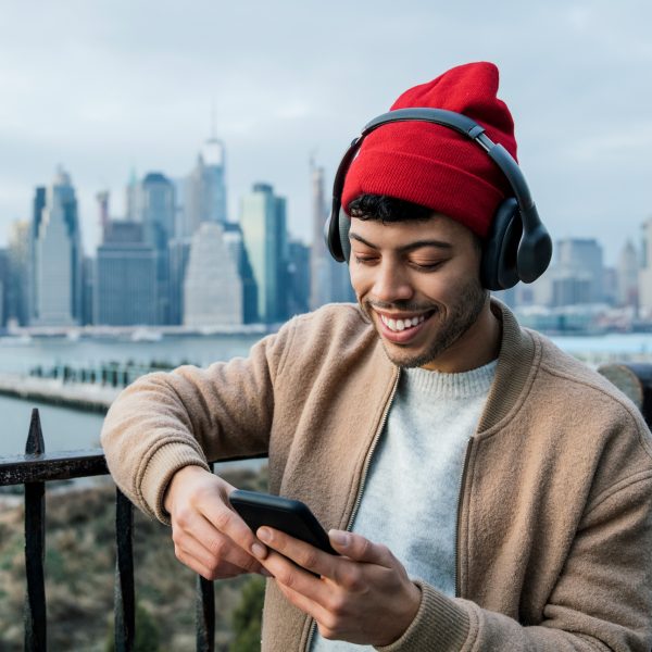 Eine Person steht vor einem Stadtpanorama und schaut auf ihr Handy, während sie über Headphones Musik hört.