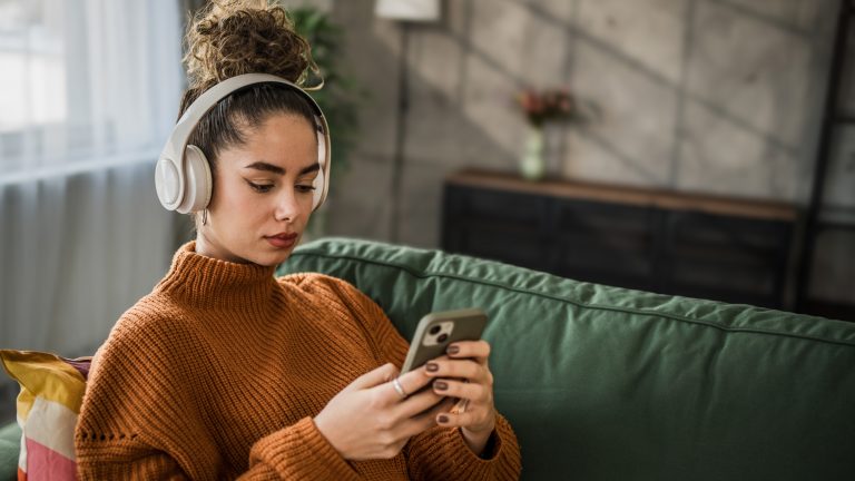 Eine Person sitzt im Wohnzimmer auf der Couch, hat Headphones auf und schaut auf ihr Smartphone.