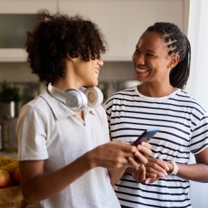 Eine erwachsene und eine jugendliche Person stehen nebeneinander und lachen sich an. Die jugendliche Person hält ein Smartphone in der Hand.