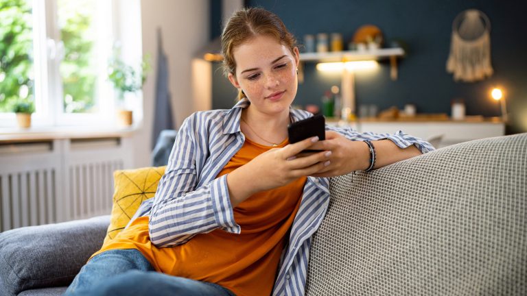 Eine jugendliche Person sitzt auf einem Sofa und hält ein Smartphone in der Hand.