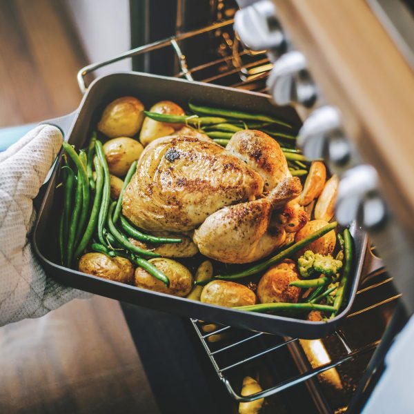 Eine Person steckt eine Auflaufform mit Hähnchen, Kartoffeln und grünen Bohnen in einen geöffneten Backofen.