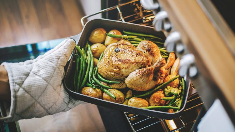 Eine Person steckt eine Auflaufform mit Hähnchen, Kartoffeln und grünen Bohnen in einen geöffneten Backofen.