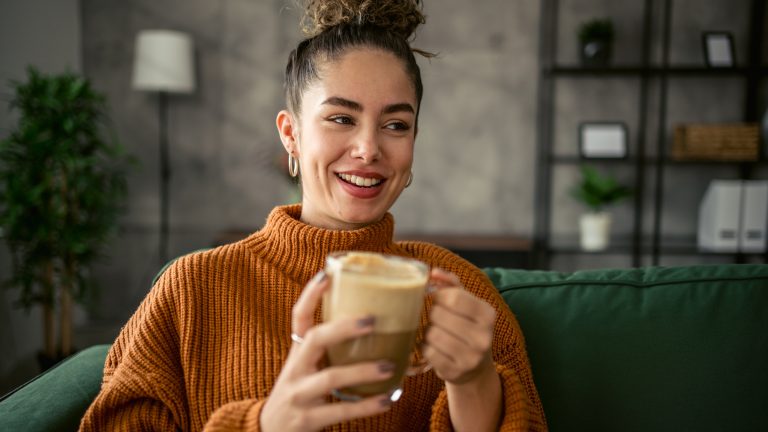 Eine Person hält eine Glastasse mit Kaffee und Milchschaum in den Händen.