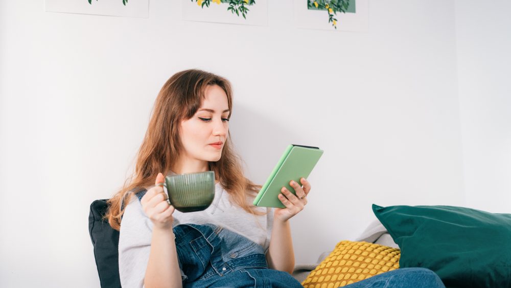 Eine Person hält eine Tasse in der Hand, während sie auf einem E Book-Reader liest.