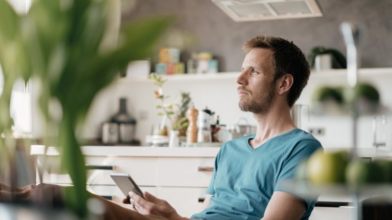 Eine Person sitzt am Tisch mit einem Smartphone und blickt nachdenklich in die Ferne.