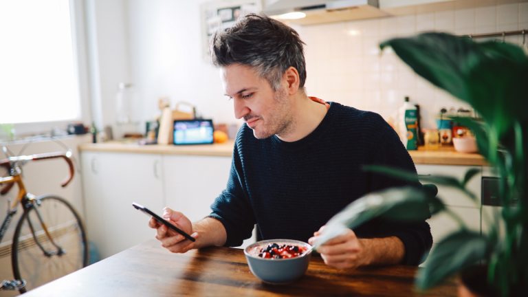 Eine Person sitzt am Küchentisch mit einer Schüssel Müsli und schaut ins Smartphone.