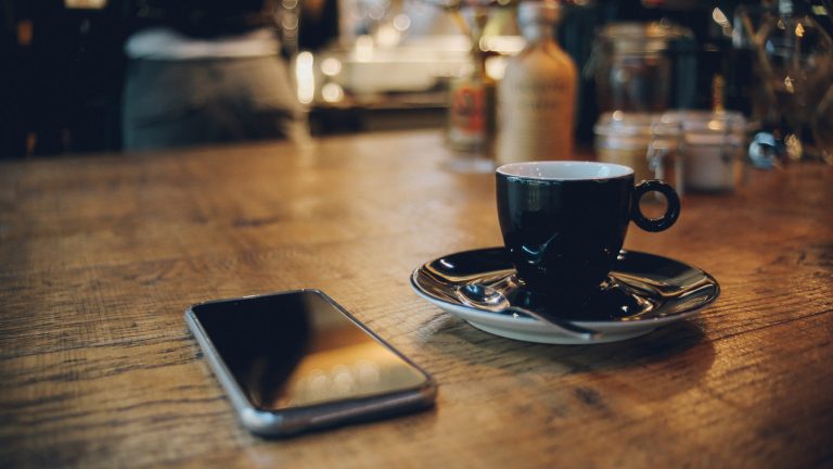 Ein Smartphone liegt auf einem Holztisch neben einer Tasse Kaffee.