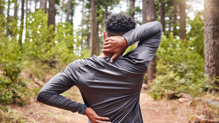 Eine Person steht in Sportkleidung im Wald und fasst sich in den Nacken.