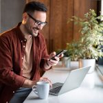 Eine Person sitzt mit einem Kaffee vor einem Laptop und hält ein Smartphone in der Hand.