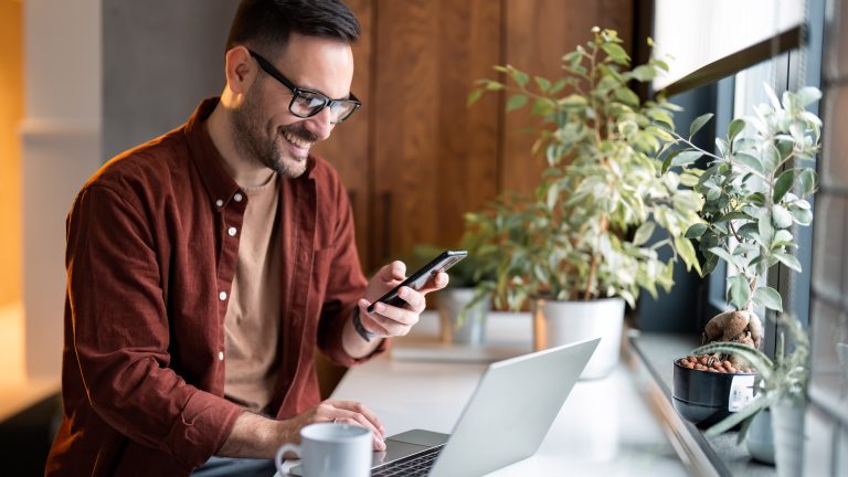 Eine Person sitzt mit einem Kaffee vor einem Laptop und hält ein Smartphone in der Hand.