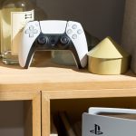 Ein Dualsense-Controller steht in einem Regal. Im Vordergrund ist eine PS5 zu erkennen.