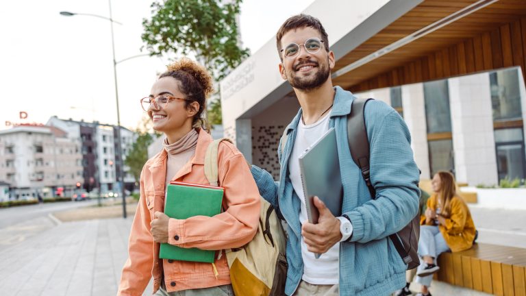 Zwei Personen entfernen sich von der Uni und tragen Arbeitsmaterial in den Händen.