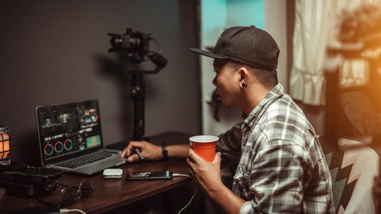 Eine Person mit einem Becher in der Hand sitzt vor einem Laptop, auf dem Videobearbeitung zu sehen ist.