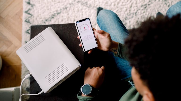 Eine Person sitzt neben einem WLAN Router und kontrolliert auf ihrem Handy die Geschwindigkeit.
