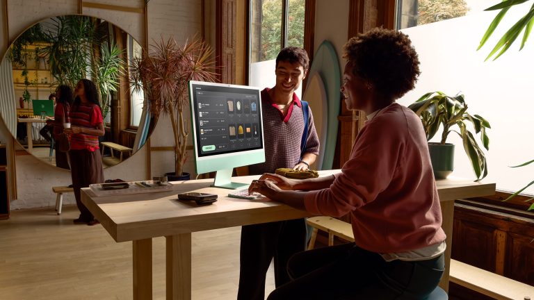 Eine Person sitzt vor einem iMac 2024. Ihr gegenüber steht eine weitere Person.