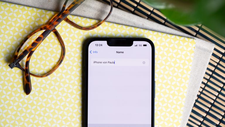 Ein iPhone liegt neben einer Brille auf einem Tisch. Auf dem Display ist das Ändern des Gerätenamens zu sehen.