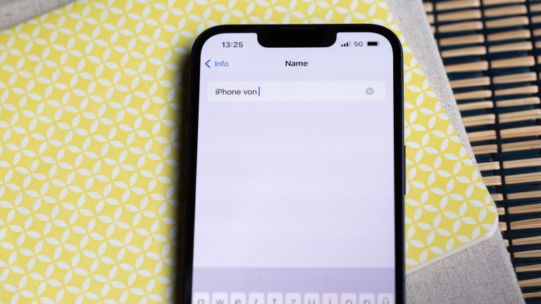 Ein iPhone liegt auf einer gelb-weißen Oberfläche. Auf dem Display ist die Namensänderung des Geräts zu sehen