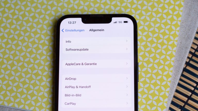 Ein iPhone liegt auf einer gelb-weißen Oberfläche. Auf dem Display sind die Allgemeinen Einstellungen zu sehen
