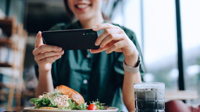 m Vordergrund steht ein Teller mit Essen, im Hintergrund ist eine Person mit Smartphone, die diesen fotografiert.