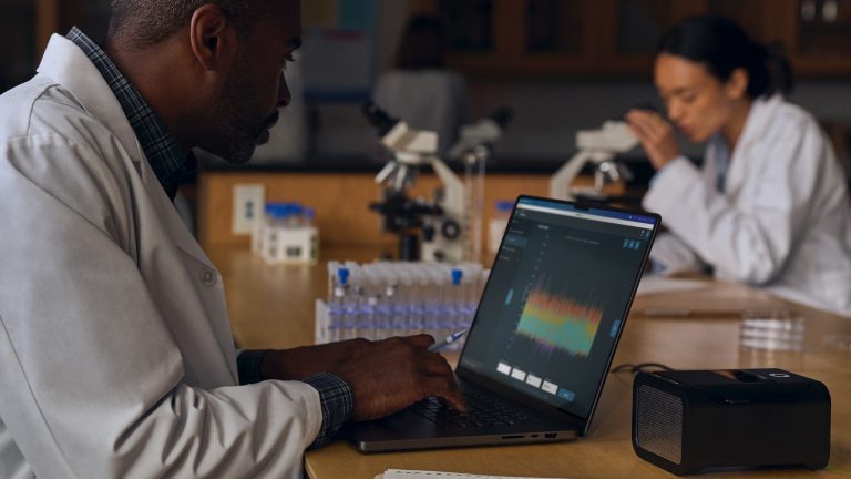 Eine Person in einem Kittel sitzt mit einem MacBook Pro in einem Labor.