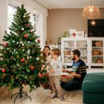 Zwei Personen sitzen auf dem Boden im Wohnzimmer, während ein Kind den Weihnachtsbaum schmückt.