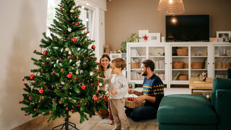 Zwei Personen sitzen auf dem Boden im Wohnzimmer, während ein Kind den Weihnachtsbaum schmückt.