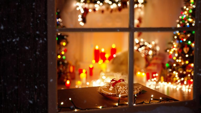 Der Blick durch ein Fenster in ein Wohnzimmer mit vielen bunten LED-Lichterketten an Weihnachtsbaum und auf Weihnachtskranz.