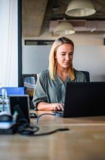 Eine Person sitzt an einem Schreibtisch nah an einem großen Fenster mit Blick auf ein Hochhaus und schaut auf einen Laptop.