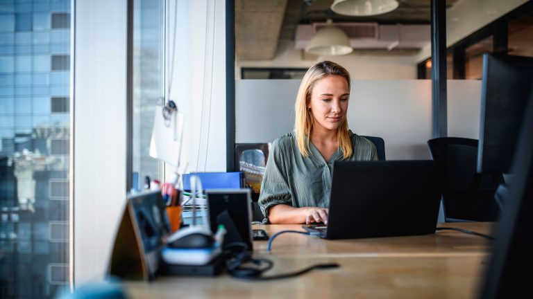 Eine Person sitzt an einem Schreibtisch nah an einem großen Fenster mit Blick auf ein Hochhaus und schaut auf einen Laptop.