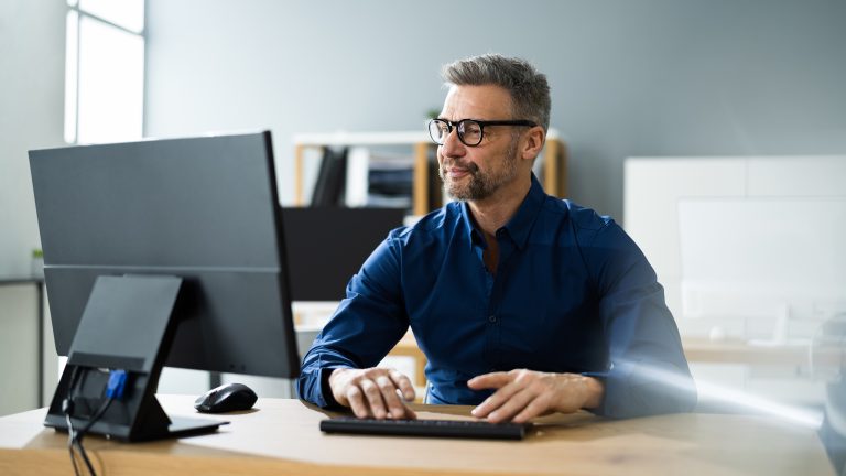 Eine Person von vorn hinter einem großen Bildschirm an einem Schreibtisch in einem cleanen Office.