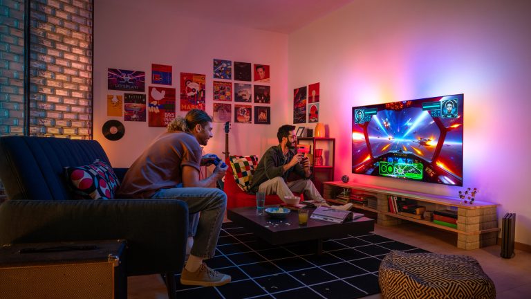 Zwei Personen sitzen vor dem Fernseher und spielen ein Videospiel. Die Wand hinter dem Fernseher ist bunt beleuchtet.