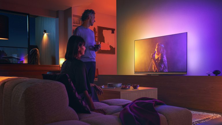 Zwei Personen betrachten einen Fernseher in einem Wohnzimmer - eine stehend, eine sitzend. Die Wand hinter dem Fernseher ist durch ihn beleuchtet.