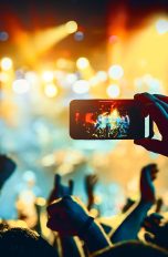 Eine Person hält ein Smartphone horizontal in die Höhe, um ein Foto von einer Bühne zu schießen auf der ein Konzert stattfindet.