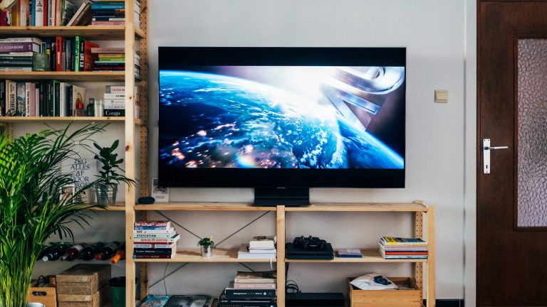 Ein Fernseher steht auf einem Holz-Lowboard in einem Wohnzimmer. Es beginnt gerade ein Spielfilm.