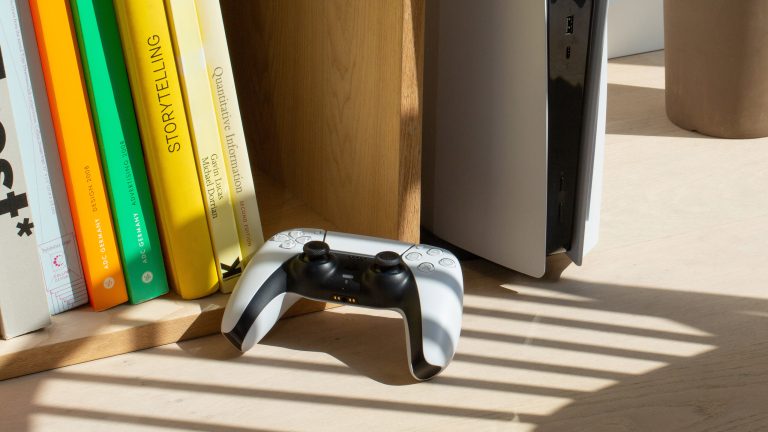 Eine PlayStation steht aufrecht in einem Bücherregal mit Büchern und davor liegt ein passender Controller.