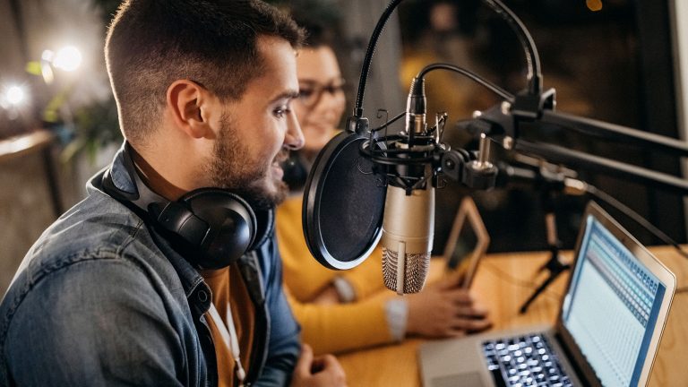 Zwei Personen sitzen vor einem Podcastmikrofon an einem Mikrofonarm und blicken dabei auf einen Laptop mit Audiosoftware.